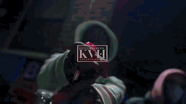 a person in a hoodie holds a red object in front of a sign that says kvh