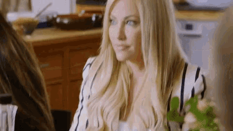 a woman with long blonde hair is sitting at a table in a kitchen with other women .