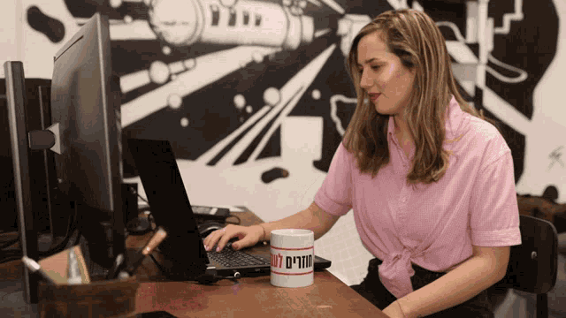 a woman in a pink shirt sits at a desk with a laptop and a mug that says ' a ' on it