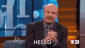 a man in a suit and tie is sitting on a stage with his hands folded and saying hello .