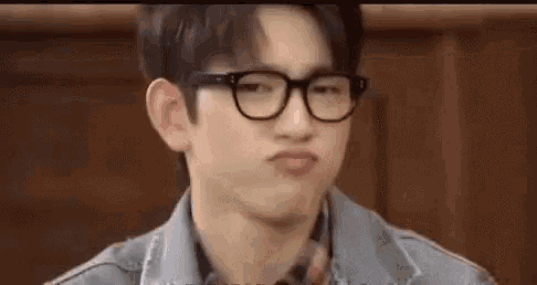 a young man wearing glasses and a denim jacket is making a face .