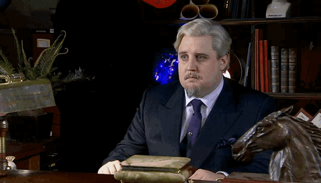 a man in a suit and tie sits at a desk with a statue of a horse and a lamp that says punchlist