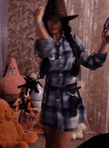 a woman wearing a plaid shirt and a cowboy hat stands in front of stuffed animals