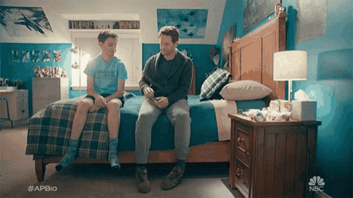 a man and a boy are sitting on a bed in a bedroom with a nbc logo on the corner