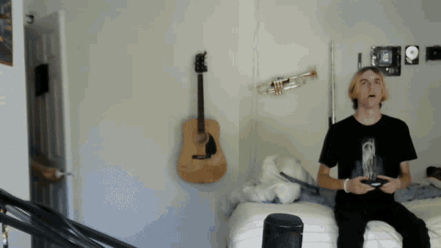 a man sitting on a bed playing a video game with a guitar and trumpet hanging on the wall behind him