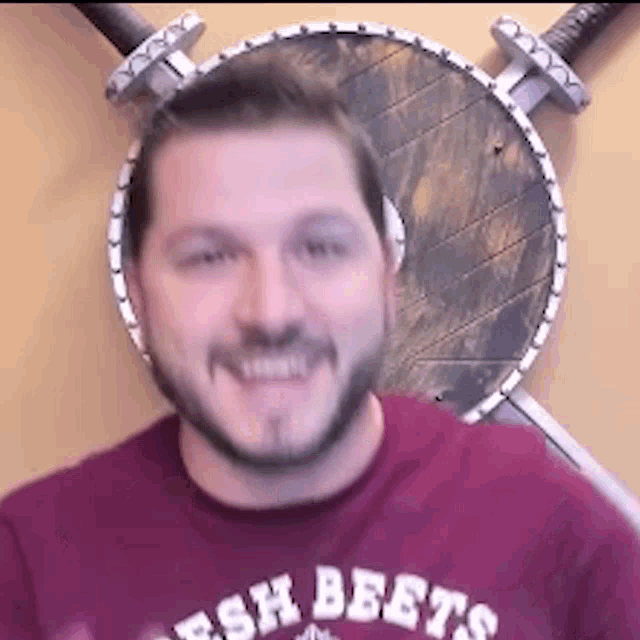 a man wearing a red shirt with the word beets on it is smiling in front of two swords and a shield .
