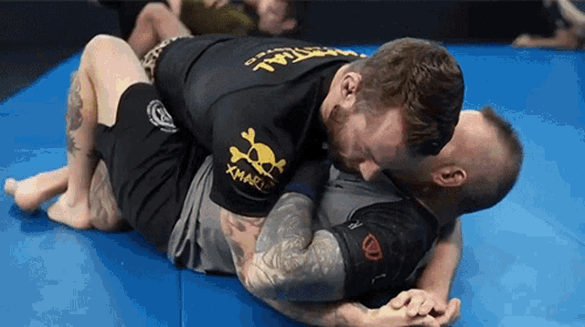 two men are wrestling on a blue mat and one of them is wearing a black shirt with a skull and crossbones on it .