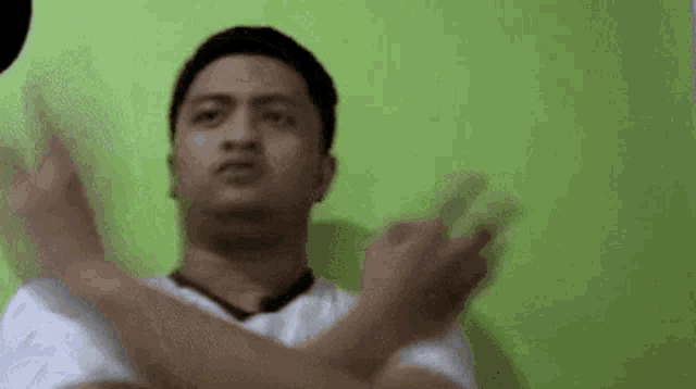 a man in a white shirt is making a funny face with his arms crossed in front of a green wall .