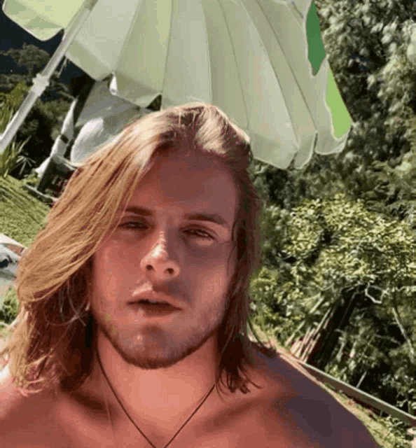 a shirtless man with long blonde hair is standing under a green umbrella