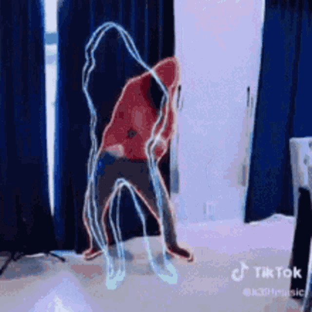 a man in a red hoodie is dancing in a room with blue curtains behind him .