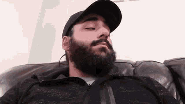 a man with a beard is sitting on a couch wearing a baseball cap .