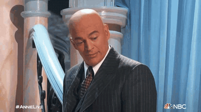 a bald man in a suit and tie stands in front of a blue curtain with the nbc logo on it