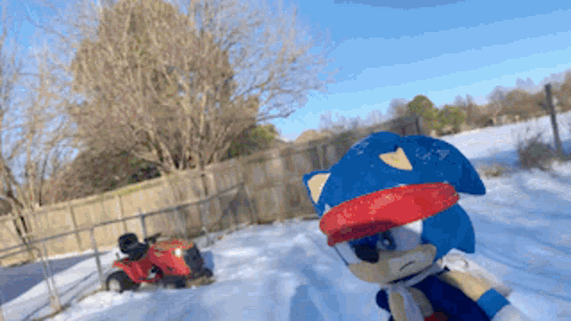 a stuffed animal of sonic the hedgehog standing in the snow with a lawn mower in the background