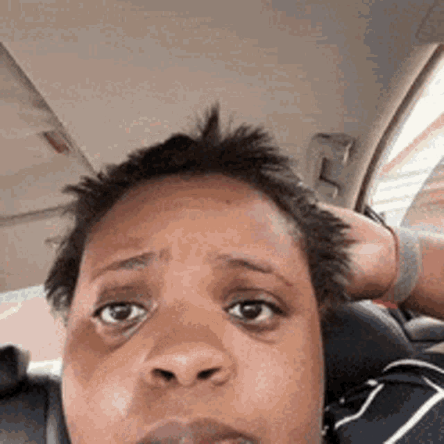 a woman is sitting in a car with her hand on her head and looking at the camera .