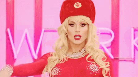 a blonde drag queen wearing a red hat and a red dress is standing in front of a pink neon sign .