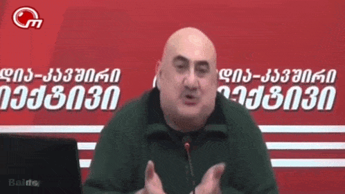bald man speaking into a microphone in front of a baldo sign