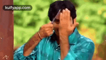 a man in a blue shirt is covering his face with his hands while holding a glass .