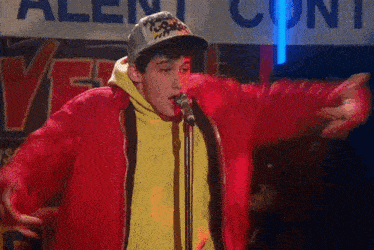 a young man is singing into a microphone while wearing a red jacket and a hat .
