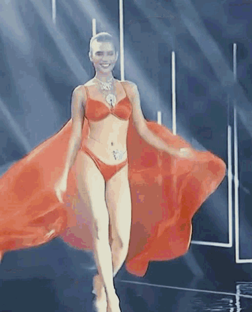 a woman in a red bikini is walking down a runway with a red cape .