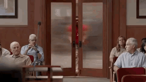 a group of people are sitting in a courtroom with a man standing in the doorway behind them