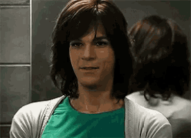 a woman wearing a wig and a green shirt is looking at her reflection in a mirror .