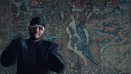 a man wearing sunglasses and a beanie is standing in front of a large tapestry