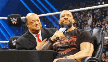 a wrestler is sitting in a chair talking into a microphone while another wrestler stands behind him .