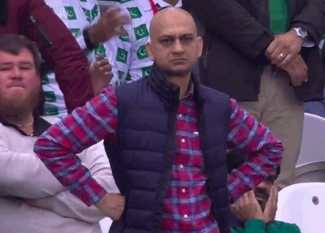 a bald man in a plaid shirt stands with his hands on his hips in a crowd