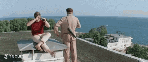 a man sitting on a roof taking a picture of the ocean while another man stands behind him