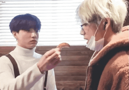 a man wearing a mask is feeding another man a piece of food while they are standing next to each other .