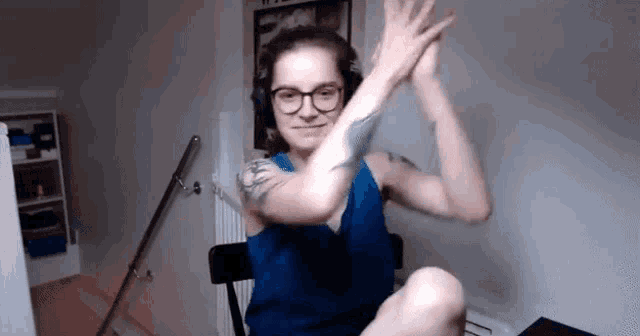 a woman wearing glasses and a blue tank top is sitting in a chair with her legs crossed and giving a high five .