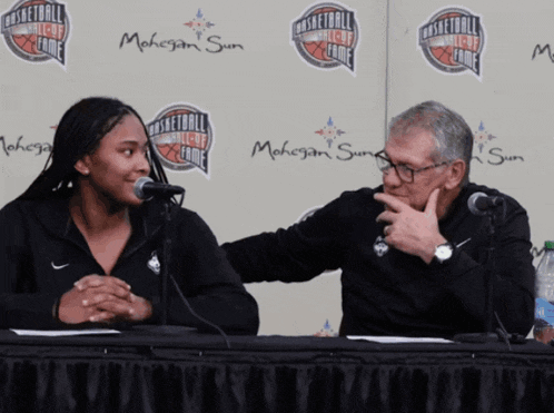 a man and a woman are sitting at a table with microphones in front of a mahogany sun sign
