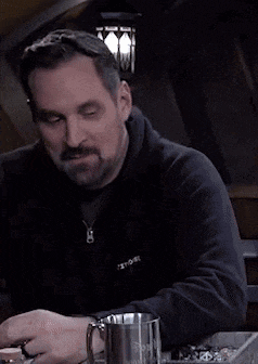 a man with a beard is sitting at a table holding a mug .