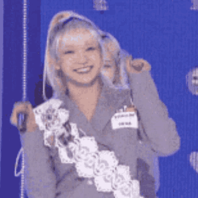 a woman with blonde hair and a name tag on her shoulder is standing in front of a blue background and smiling .