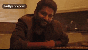 a man with a beard is sitting at a table in a kitchen talking to someone .