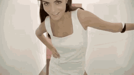 a woman in a white tank top is standing with her arms outstretched and looking at the camera .