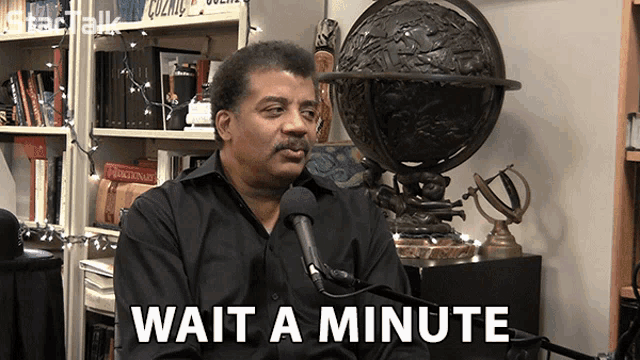 a man sitting in front of a microphone with wait a minute written on the bottom