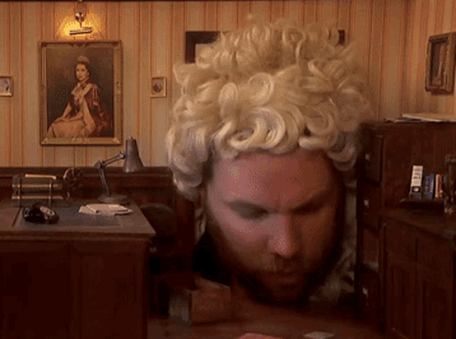 a man with a wig on his head is sitting at a desk in front of a painting of a woman