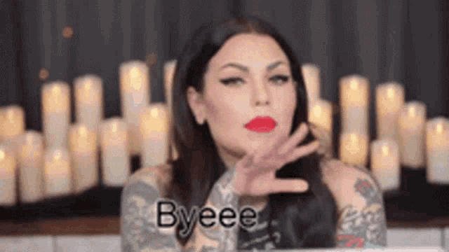 a woman with a tattoo on her arm is standing in front of a bunch of candles and saying byeee .