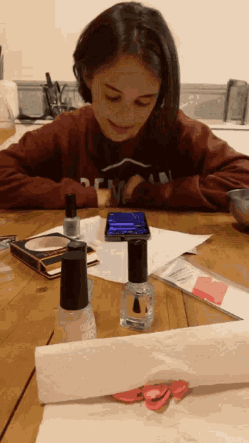 a girl is sitting at a table with bottles of nail polish on it