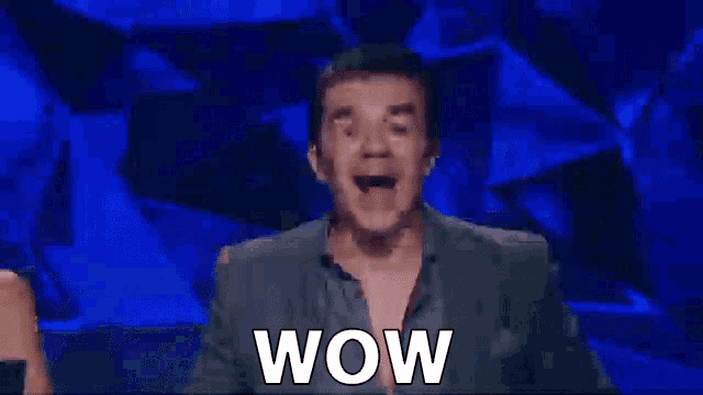 a man in a suit is sitting on a stage with his mouth open and the word wow written on the screen .