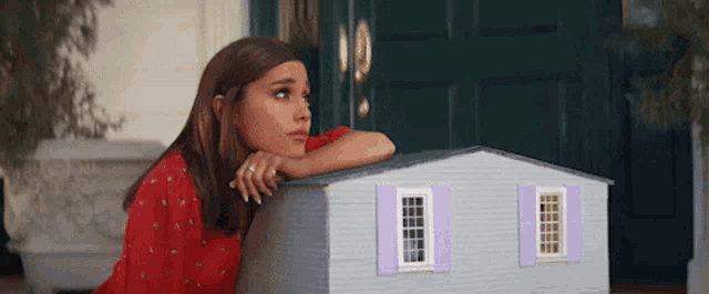 a woman in a red dress leans against a small doll house