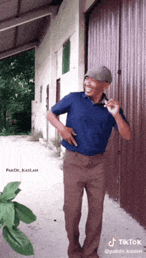 a man wearing a blue shirt and brown pants is standing in front of a building with a tiktok logo on the bottom right