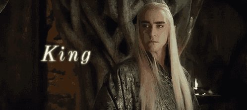 a man with long white hair and a crown on his head is standing in front of a tree with the word king above him .