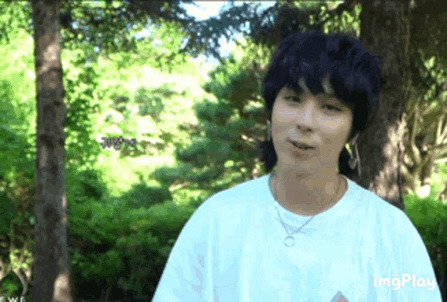 a young man wearing a white t-shirt and a necklace stands in front of trees