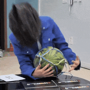 a woman in a blue jacket is holding a watermelon on a table with chanel 9 cards on it