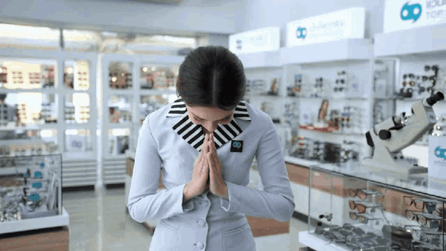 a woman wearing a white jacket with a black and white striped collar is praying in front of a display of glasses