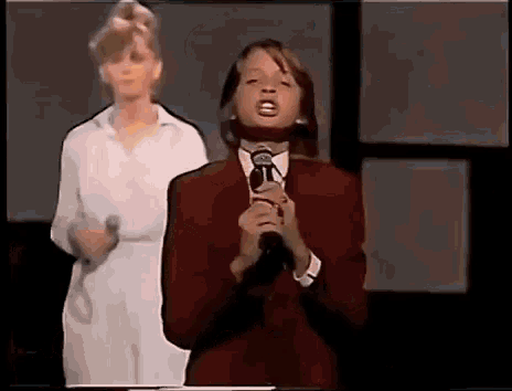 a young man in a red suit is singing into a microphone while a woman stands behind him .