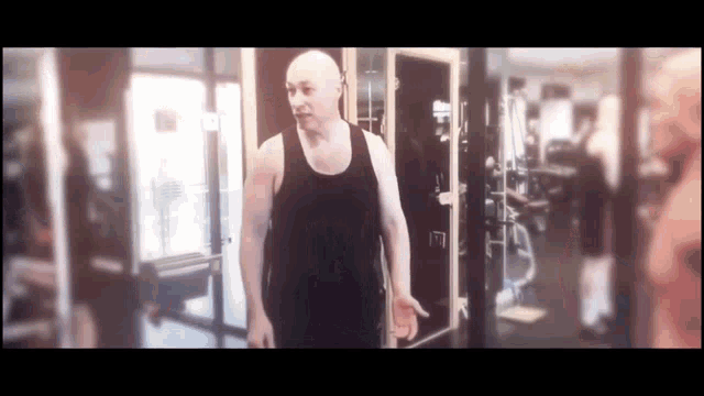 a bald man in a black tank top is walking through a gym .