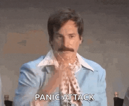a man with a mustache is sitting at a table with his hands folded in front of his face and says panic attack .